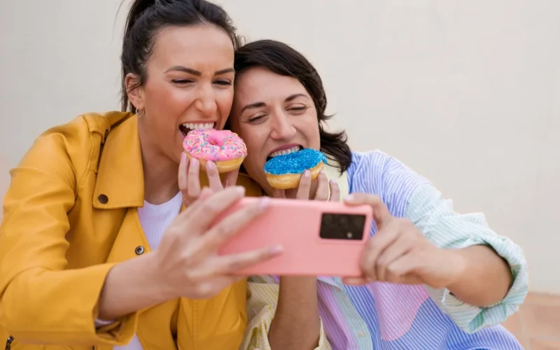 Two women smiling while holding colorful donuts and taking a selfie with a smartphone.